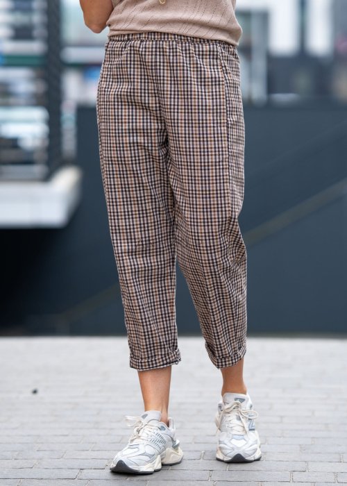 Beżowo-brązowe spodnie damskie w drobną kratę o długości 7/8 z elastyczną talią i kieszeniami, zestawione ze sneakersami w miejskiej stylizacji.