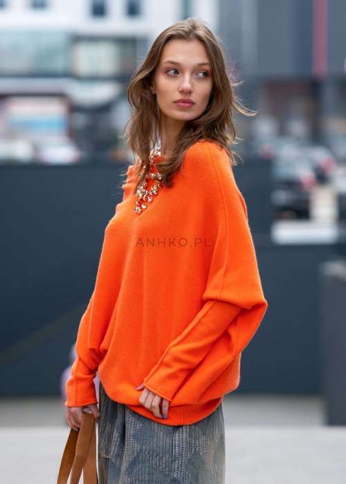 Pomarańczowy sweter damski oversize o nietoperzowym kroju z asymetrycznym dołem, stylizacja miejska z biżuterią i spodniami.