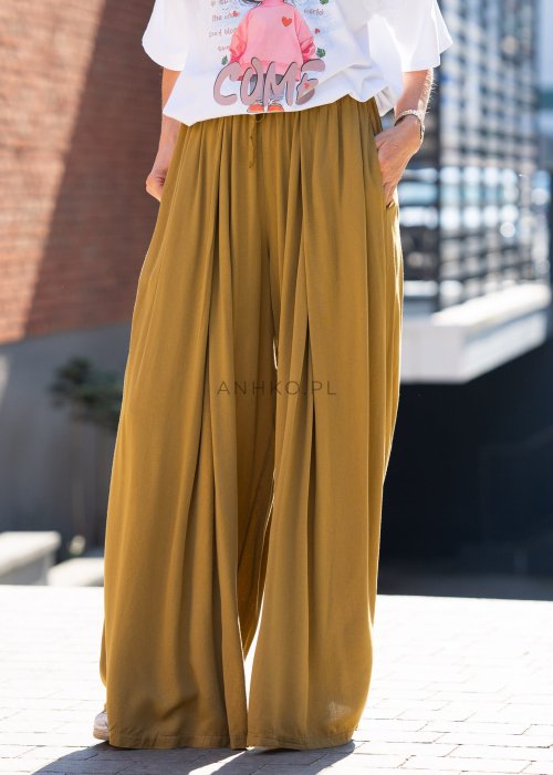 Musztardowe spodnie damskie z szeroką, lejącą nogawką i elastycznym pasem, stylizowane z białym t-shirtem w casualowym, miejskim stylu.