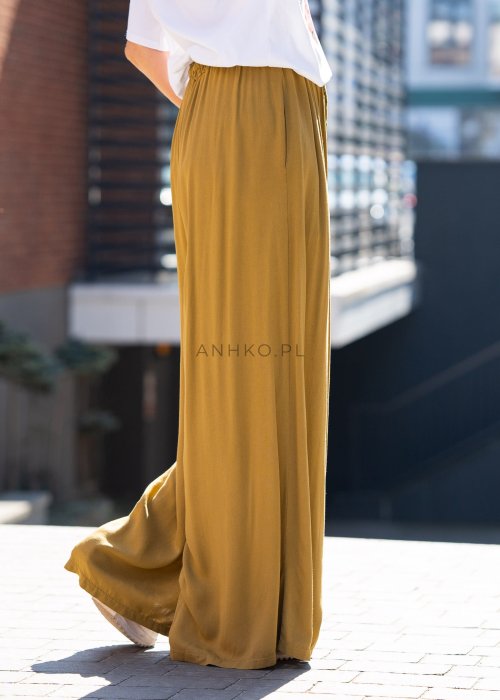 Musztardowe spodnie damskie z szeroką, lejącą nogawką i elastycznym pasem, stylizowane z białym t-shirtem w casualowym, miejskim stylu.
