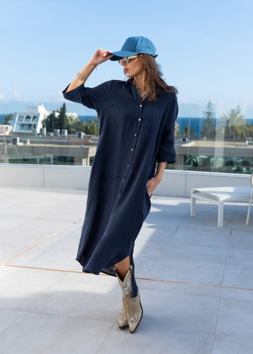 Granatowa lniana sukienka maxi w stylu szmizjerki, zapinana na guziki, o luźnym fasonie oversize, stylizowana z kowbojkami i czapką.