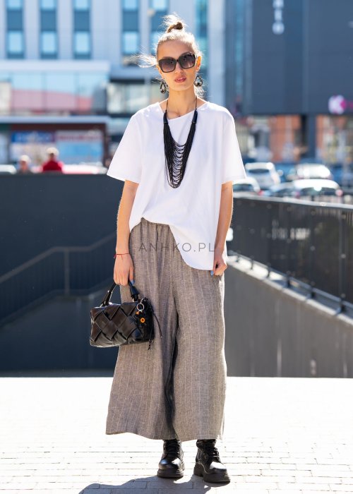 Biała bluzka damska o luźnym, asymetrycznym kroju z krótkim rękawem. Model prezentowany w stylizacji z szerokimi spodniami, podkreślającej nowoczesny i minimalistyczny charakter fasonu.