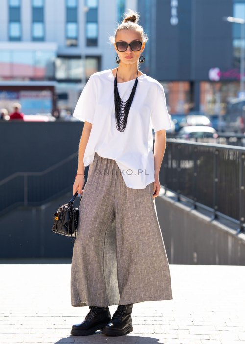 Biała bluzka damska o luźnym, asymetrycznym kroju z krótkim rękawem. Model prezentowany w stylizacji z szerokimi spodniami, podkreślającej nowoczesny i minimalistyczny charakter fasonu.