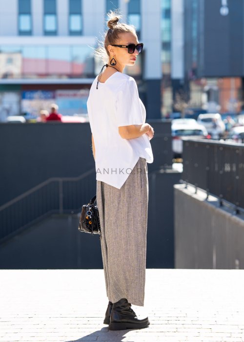 Biała bluzka damska o luźnym, asymetrycznym kroju z krótkim rękawem. Model prezentowany w stylizacji z szerokimi spodniami, podkreślającej nowoczesny i minimalistyczny charakter fasonu.