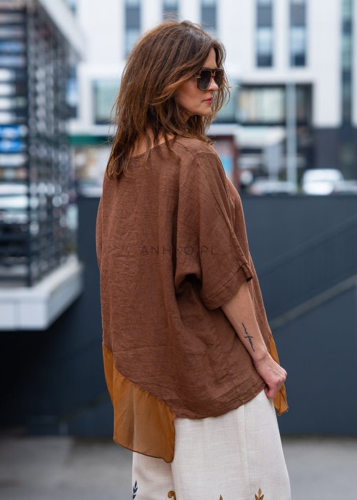 Brązowa bluzka lniana oversize z asymetrycznym dołem i lekką, transparentną wstawką, zestawiona z jasnymi spodniami w miejskiej stylizacji.