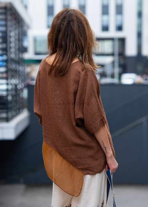 Brązowa bluzka lniana oversize z asymetrycznym dołem i lekką, transparentną wstawką, zestawiona z jasnymi spodniami w miejskiej stylizacji.