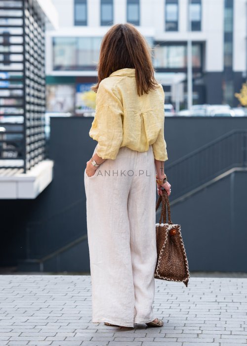 Kremowe lniane spodnie damskie o szerokiej nogawce z elastycznym pasem i ozdobnym paskiem boho, stylizowane z żółtą lnianą koszulą i torebką plecioną.