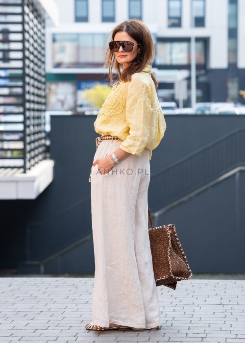 Kremowe lniane spodnie damskie o szerokiej nogawce z elastycznym pasem i ozdobnym paskiem boho, stylizowane z żółtą lnianą koszulą i torebką plecioną.