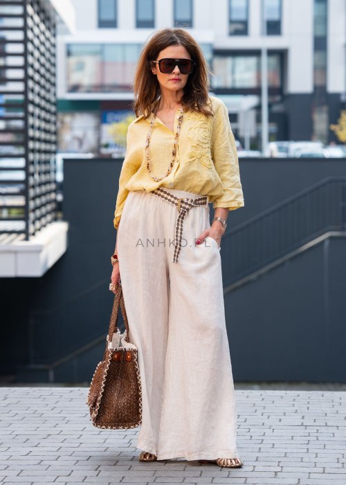Kremowe lniane spodnie damskie o szerokiej nogawce z elastycznym pasem i ozdobnym paskiem boho, stylizowane z żółtą lnianą koszulą i torebką plecioną.