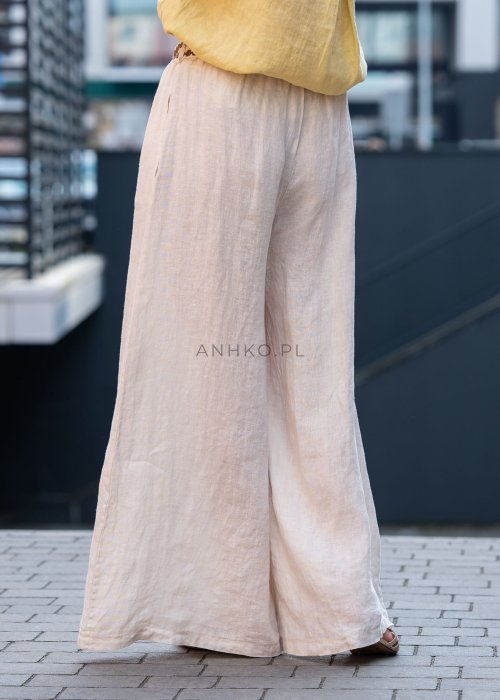 Kremowe lniane spodnie damskie o szerokiej nogawce z elastycznym pasem i ozdobnym paskiem boho, stylizowane z żółtą lnianą koszulą i torebką plecioną.