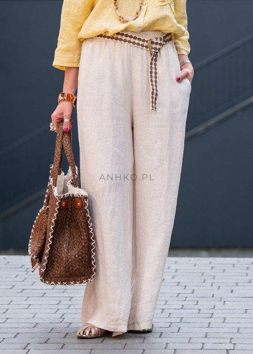 Kremowe lniane spodnie damskie o szerokiej nogawce z elastycznym pasem i ozdobnym paskiem boho, stylizowane z żółtą lnianą koszulą i torebką plecioną.