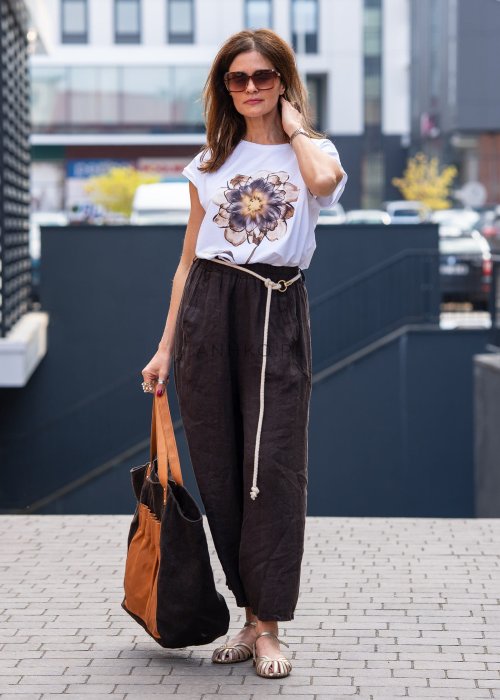 Ciemnobrązowe lniane spodnie damskie z szeroką nogawką i elastycznym pasem ze sznurkiem. Stylizacja casual z białą bluzką i sandałami.