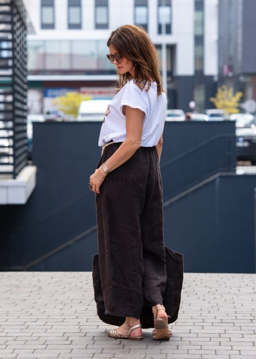 Ciemnobrązowe lniane spodnie damskie z szeroką nogawką i elastycznym pasem ze sznurkiem. Stylizacja casual z białą bluzką i sandałami.