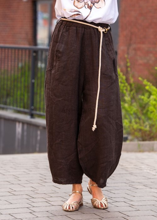 Ciemnobrązowe lniane spodnie damskie z szeroką nogawką i elastycznym pasem ze sznurkiem. Stylizacja casual z białą bluzką i sandałami.
