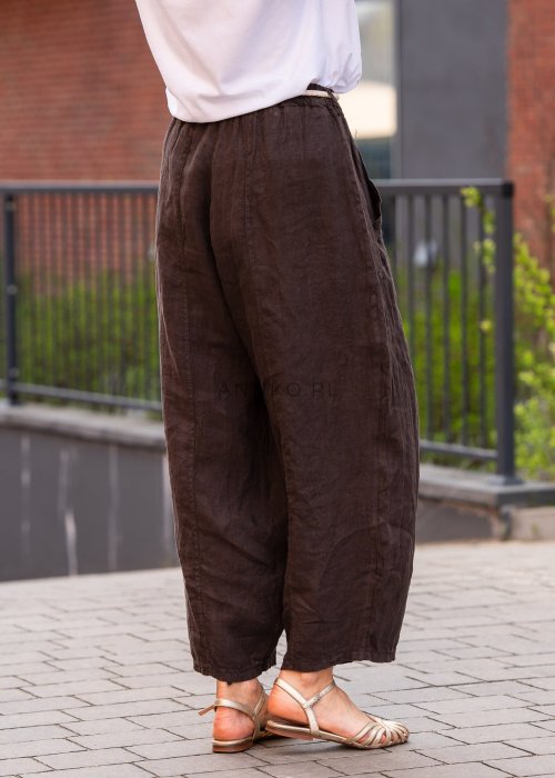 Ciemnobrązowe lniane spodnie damskie z szeroką nogawką i elastycznym pasem ze sznurkiem. Stylizacja casual z białą bluzką i sandałami.