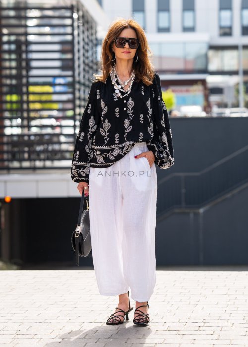 Białe lniane spodnie damskie z szeroką nogawką i elastycznym pasem ze sznurkiem, stylizowane z czarną haftowaną bluzką i sandałkami na obcasie.