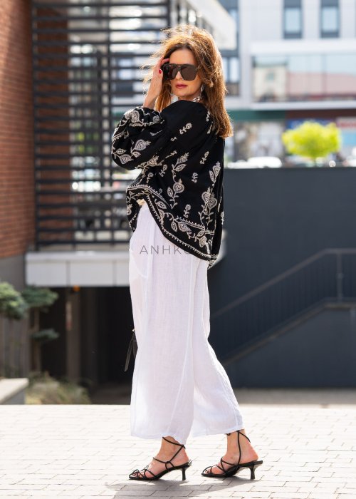 Białe lniane spodnie damskie z szeroką nogawką i elastycznym pasem ze sznurkiem, stylizowane z czarną haftowaną bluzką i sandałkami na obcasie.