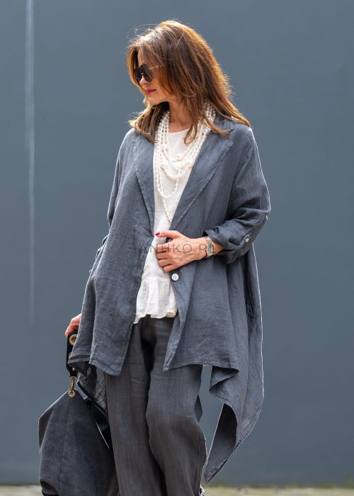 Antracytowa lniana marynarka damska o asymetrycznym kroju z klasycznym kołnierzem i podwijanymi rękawami. Model pokazany w stylizacji z jasną bluzką i lnianymi spodniami.