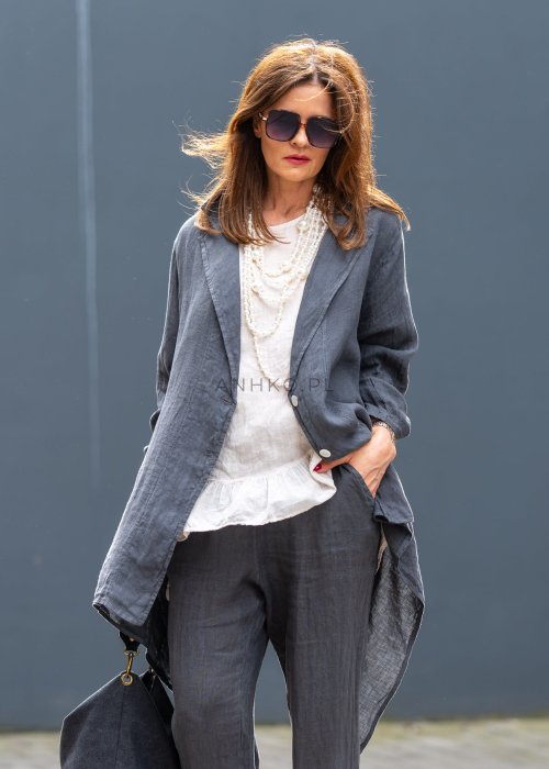 Antracytowa lniana marynarka damska o asymetrycznym kroju z klasycznym kołnierzem i podwijanymi rękawami. Model pokazany w stylizacji z jasną bluzką i lnianymi spodniami.