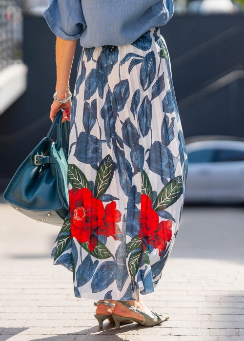 Długa spódnica lniana w kwiatowy wzór z czerwonymi akcentami i granatowymi liśćmi na jasnym tle, zestawiona z koszulą i torebką w stylu casual.