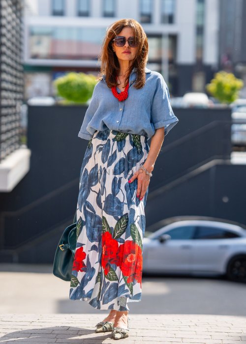 Długa spódnica lniana w kwiatowy wzór z czerwonymi akcentami i granatowymi liśćmi na jasnym tle, zestawiona z koszulą i torebką w stylu casual.
