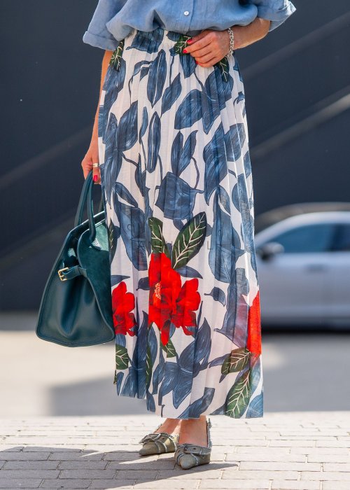 Długa spódnica lniana w kwiatowy wzór z czerwonymi akcentami i granatowymi liśćmi na jasnym tle, zestawiona z koszulą i torebką w stylu casual.