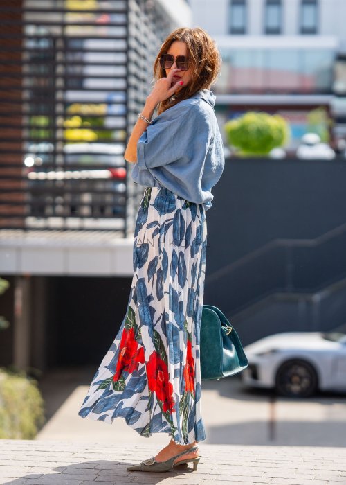 Długa spódnica lniana w kwiatowy wzór z czerwonymi akcentami i granatowymi liśćmi na jasnym tle, zestawiona z koszulą i torebką w stylu casual.