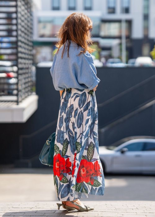 Długa spódnica lniana w kwiatowy wzór z czerwonymi akcentami i granatowymi liśćmi na jasnym tle, zestawiona z koszulą i torebką w stylu casual.