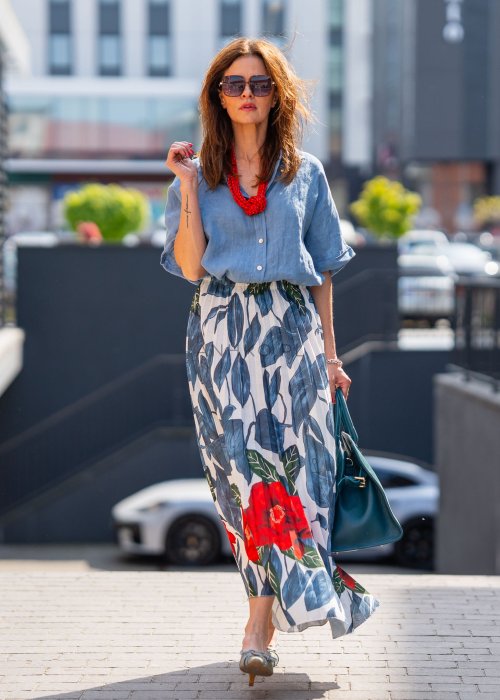 Długa spódnica lniana w kwiatowy wzór z czerwonymi akcentami i granatowymi liśćmi na jasnym tle, zestawiona z koszulą i torebką w stylu casual.