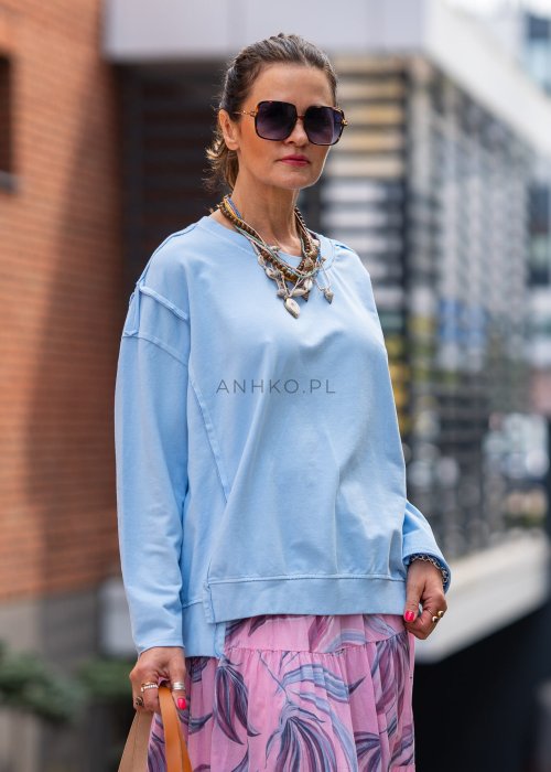 Damska bluza oversize w jasnym błękitnym kolorze z długim rękawem i okrągłym dekoltem. Stylizacja miejska zestawiona z różową spódnicą w roślinny wzór i dużymi okularami przeciwsłonecznymi.