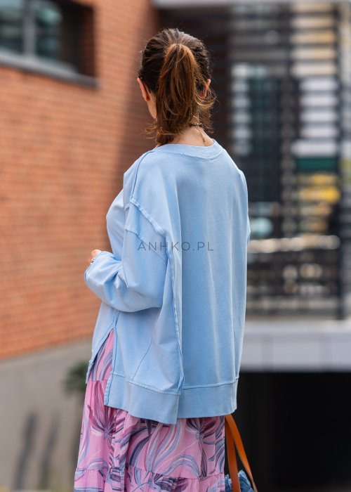 Damska bluza oversize w jasnym błękitnym kolorze z długim rękawem i okrągłym dekoltem. Stylizacja miejska zestawiona z różową spódnicą w roślinny wzór i dużymi okularami przeciwsłonecznymi.