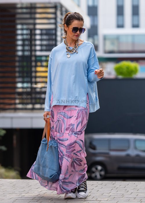 Damska bluza oversize w jasnym błękitnym kolorze z długim rękawem i okrągłym dekoltem. Stylizacja miejska zestawiona z różową spódnicą w roślinny wzór i dużymi okularami przeciwsłonecznymi.