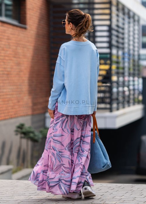 Damska bluza oversize w jasnym błękitnym kolorze z długim rękawem i okrągłym dekoltem. Stylizacja miejska zestawiona z różową spódnicą w roślinny wzór i dużymi okularami przeciwsłonecznymi.