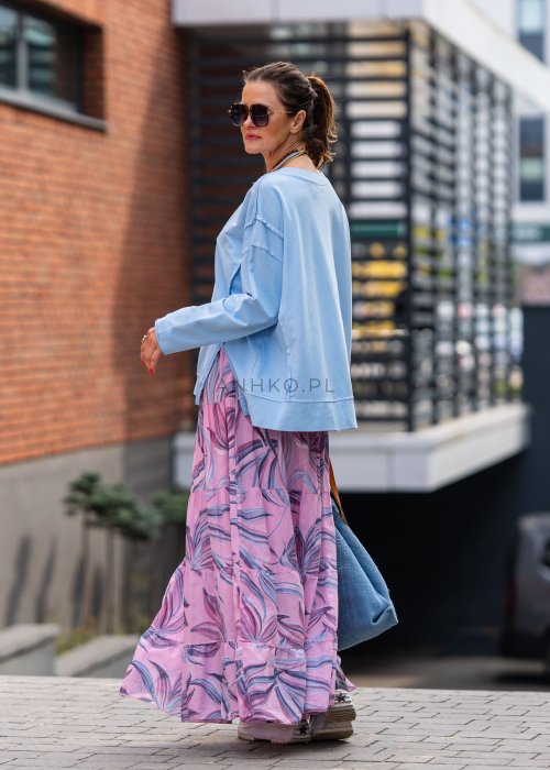 Damska bluza oversize w jasnym błękitnym kolorze z długim rękawem i okrągłym dekoltem. Stylizacja miejska zestawiona z różową spódnicą w roślinny wzór i dużymi okularami przeciwsłonecznymi.