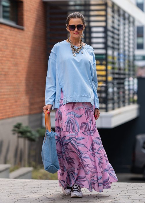 Damska bluza oversize w jasnym błękitnym kolorze z długim rękawem i okrągłym dekoltem. Stylizacja miejska zestawiona z różową spódnicą w roślinny wzór i dużymi okularami przeciwsłonecznymi.