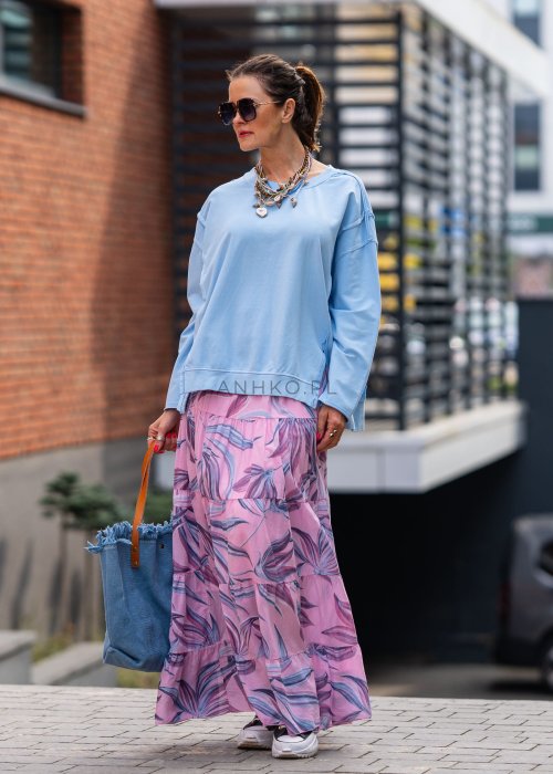 Damska bluza oversize w jasnym błękitnym kolorze z długim rękawem i okrągłym dekoltem. Stylizacja miejska zestawiona z różową spódnicą w roślinny wzór i dużymi okularami przeciwsłonecznymi.