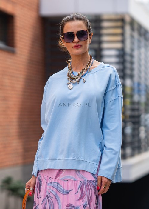 Damska bluza oversize w jasnym błękitnym kolorze z długim rękawem i okrągłym dekoltem. Stylizacja miejska zestawiona z różową spódnicą w roślinny wzór i dużymi okularami przeciwsłonecznymi.
