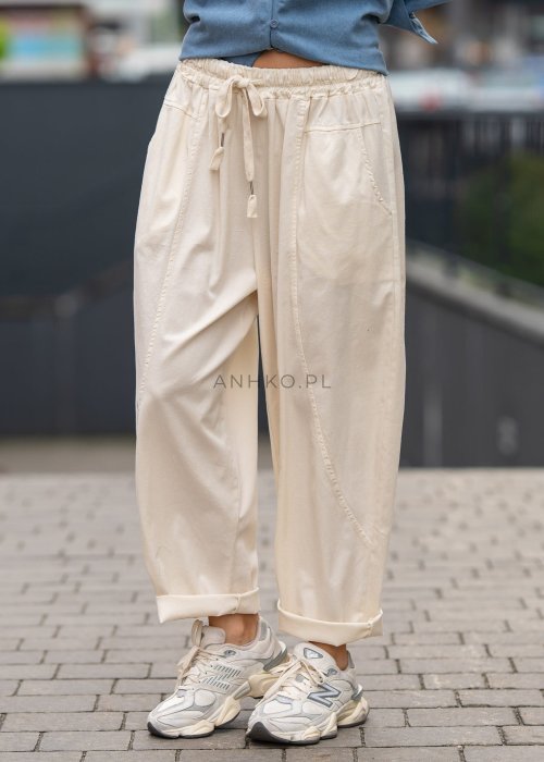 Śmietankowe spodnie damskie o luźnym kroju boyfriend z elastycznym pasem i troczkiem, zestawione ze sneakersami. Model ma podwijane nogawki i praktyczne kieszenie, tworząc swobodną stylizację miejską.