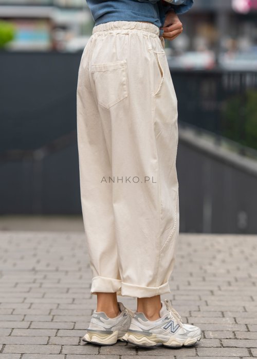 Śmietankowe spodnie damskie o luźnym kroju boyfriend z elastycznym pasem i troczkiem, zestawione ze sneakersami. Model ma podwijane nogawki i praktyczne kieszenie, tworząc swobodną stylizację miejską.