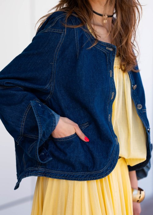 Krótka kurtka jeansowa damska bez kołnierza z ozdobnymi przeszyciami i napami, stylizowana z żółtą sukienką i biżuterią.