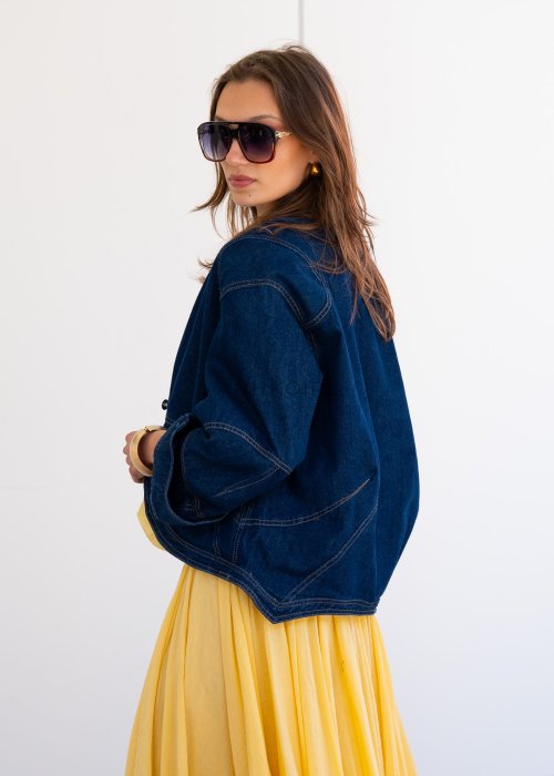 Krótka kurtka jeansowa damska bez kołnierza z ozdobnymi przeszyciami i napami, stylizowana z żółtą sukienką i biżuterią.