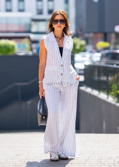 Biała kamizelka lniana damska w pionowe paski z klapami i zapięciem na dwa guziki, zestawiona z szerokimi spodniami w komplecie. Elegancka stylizacja miejska z lekką kamizelką bez rękawów.