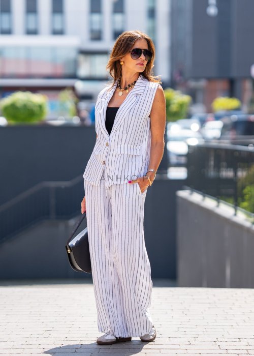 Biała kamizelka lniana damska w pionowe paski z klapami i zapięciem na dwa guziki, zestawiona z szerokimi spodniami w komplecie. Elegancka stylizacja miejska z lekką kamizelką bez rękawów.