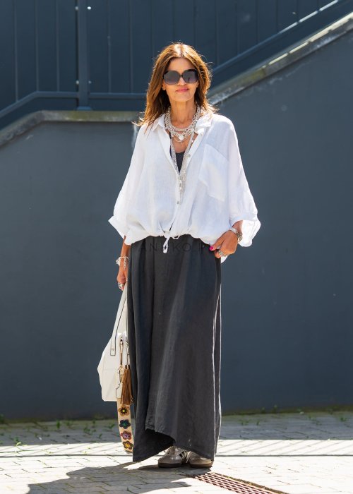 Biała koszula lniana damska o luźnym kroju z kołnierzykiem i kieszonką, zestawiona z czarną spódnicą i minimalistyczną biżuterią w eleganckim stylu.