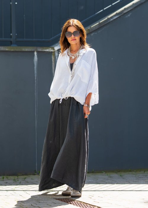 Biała koszula lniana damska o luźnym kroju z kołnierzykiem i kieszonką, zestawiona z czarną spódnicą i minimalistyczną biżuterią w eleganckim stylu.