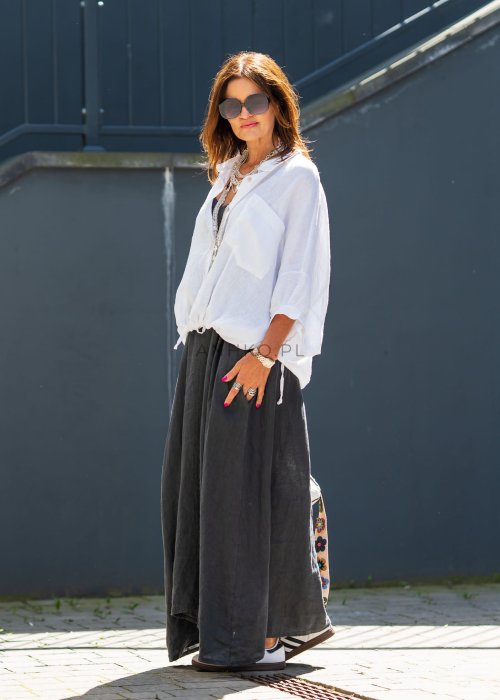 Biała koszula lniana damska o luźnym kroju z kołnierzykiem i kieszonką, zestawiona z czarną spódnicą i minimalistyczną biżuterią w eleganckim stylu.