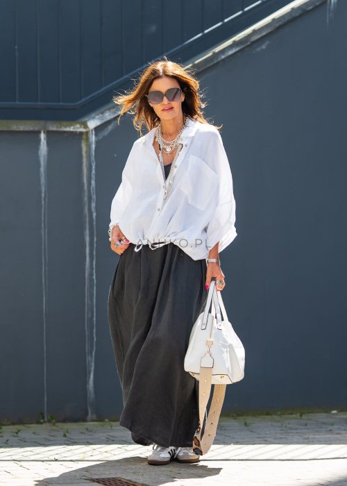 Biała koszula lniana damska o luźnym kroju z kołnierzykiem i kieszonką, zestawiona z czarną spódnicą i minimalistyczną biżuterią w eleganckim stylu.