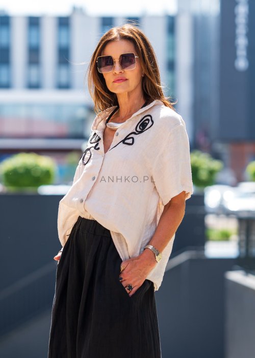 Beżowa bluzka lniana z krótkim rękawem i czarnym, artystycznym haftem na przodzie. Stylizacja z czarną spódnicą maxi i minimalistyczną biżuterią.