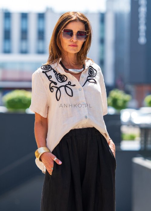 Beżowa bluzka lniana z krótkim rękawem i czarnym, artystycznym haftem na przodzie. Stylizacja z czarną spódnicą maxi i minimalistyczną biżuterią.