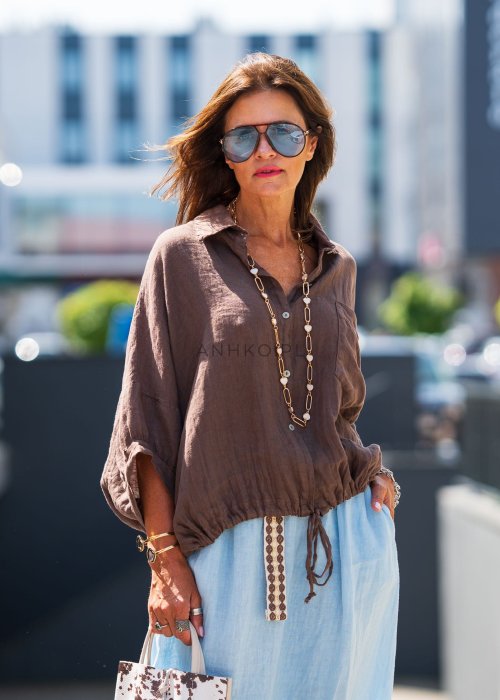 Brązowa koszula lniana damska o luźnym kroju z kołnierzykiem i kieszonką na piersi, zestawiona z jasną, lnianą spódnicą i dodatkami w naturalnym stylu.